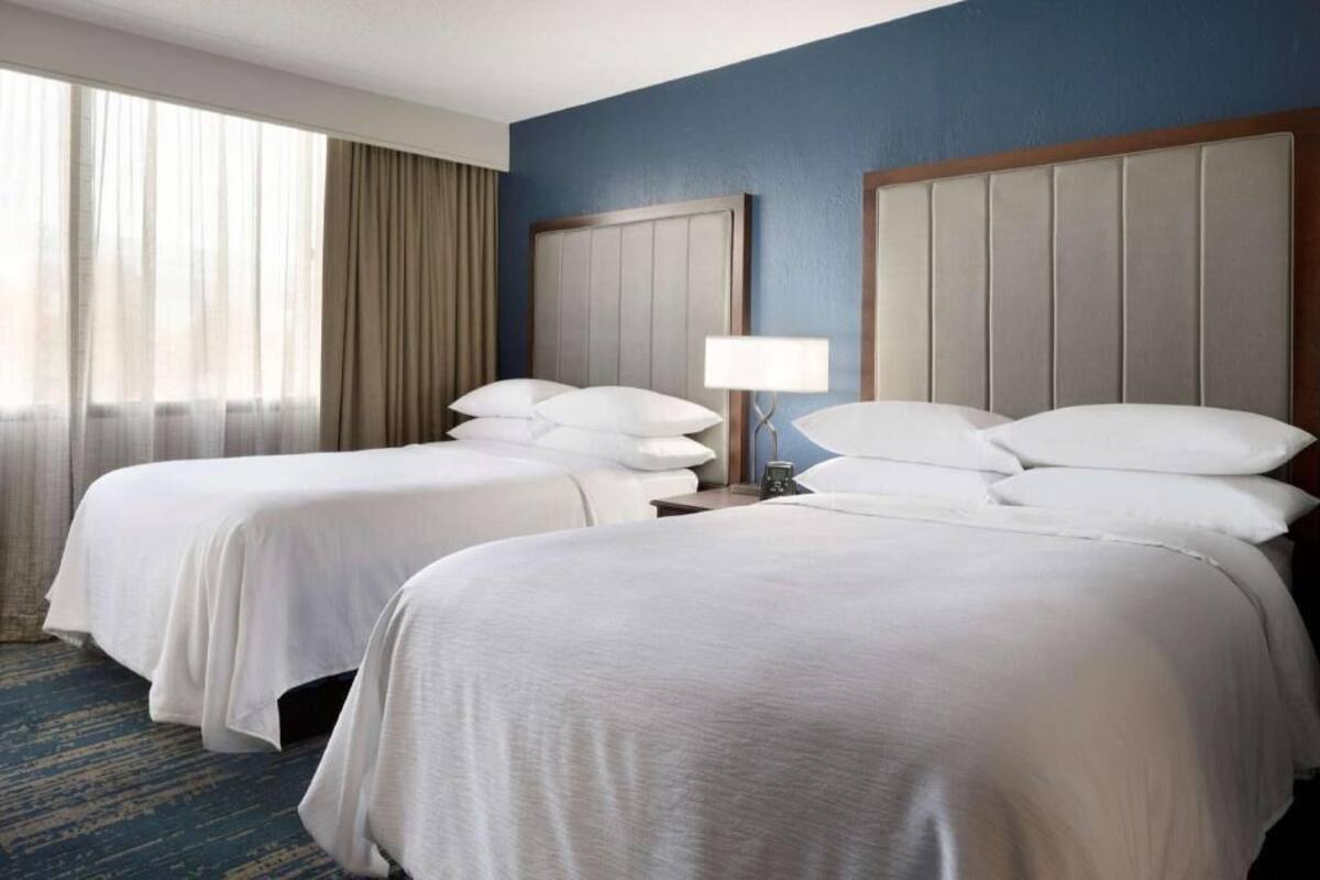 A hotel room with two beds, a nightstand, a lamp, and a large window with curtains.