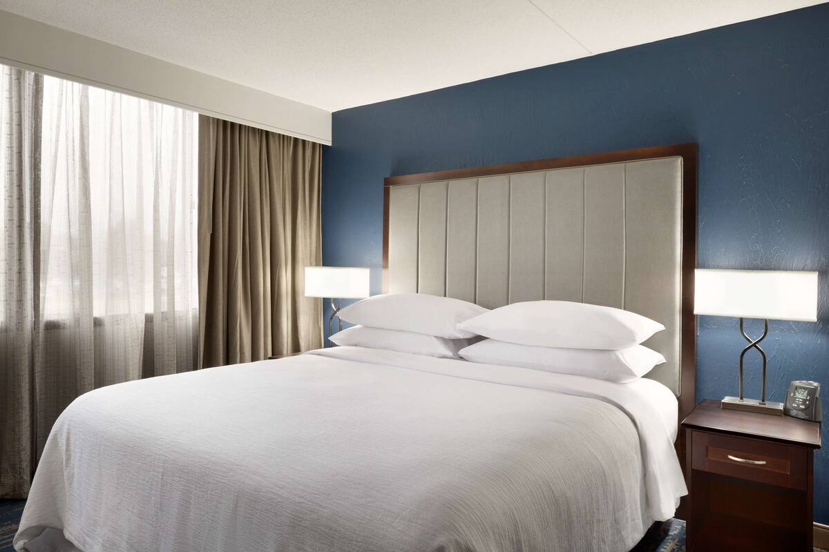A hotel room with a large bed, two bedside lamps, a nightstand, and a window with curtains.