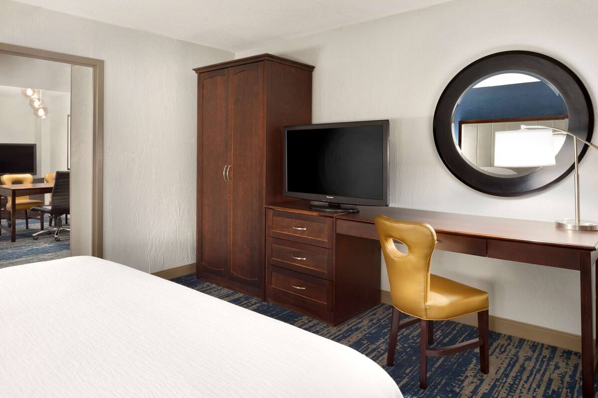A hotel room with a bed, a desk with a television, a wardrobe, a chair, and a round mirror on the wall.