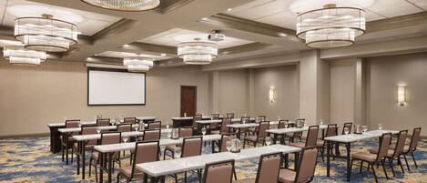 A conference room with multiple tables and chairs, large chandeliers, and a projector screen.