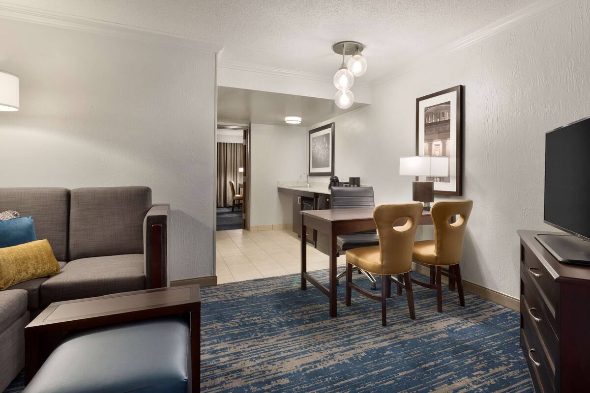 A hotel room with a living area, a dining table with chairs, a TV, and a bathroom visible in the background.
