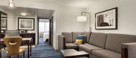 A hotel room with a sofa, a chair, a small table, a lamp, and a framed picture on the wall.