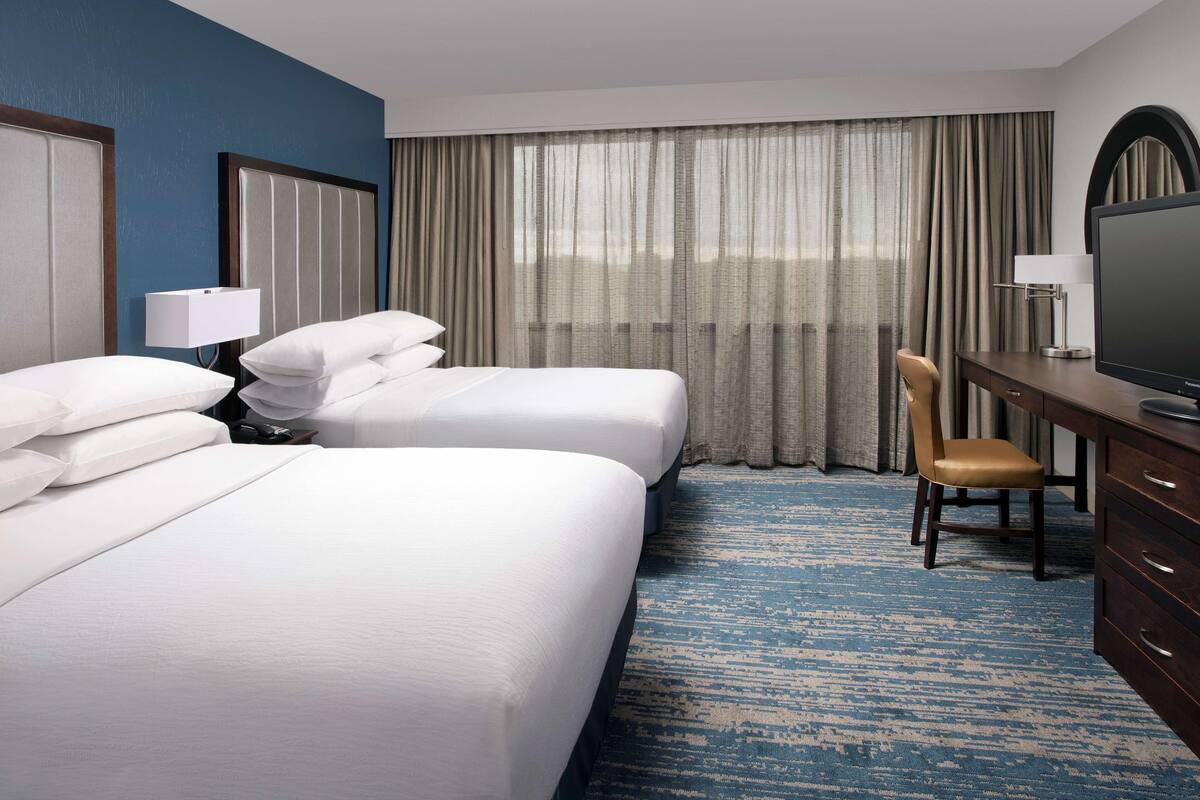 A hotel room with two beds, a desk, a chair, a television, and a large window with curtains.