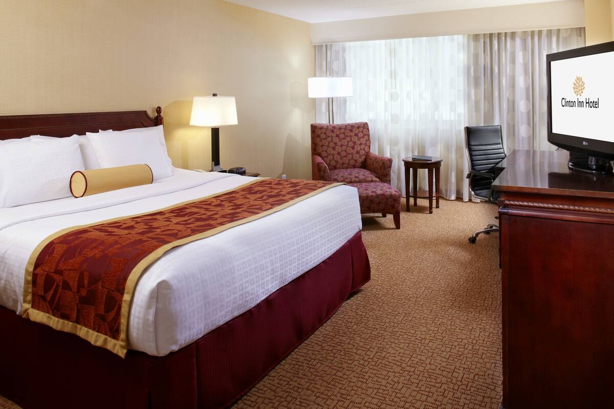 A hotel room with a large bed, two chairs, a desk, and a television displaying the hotel name.