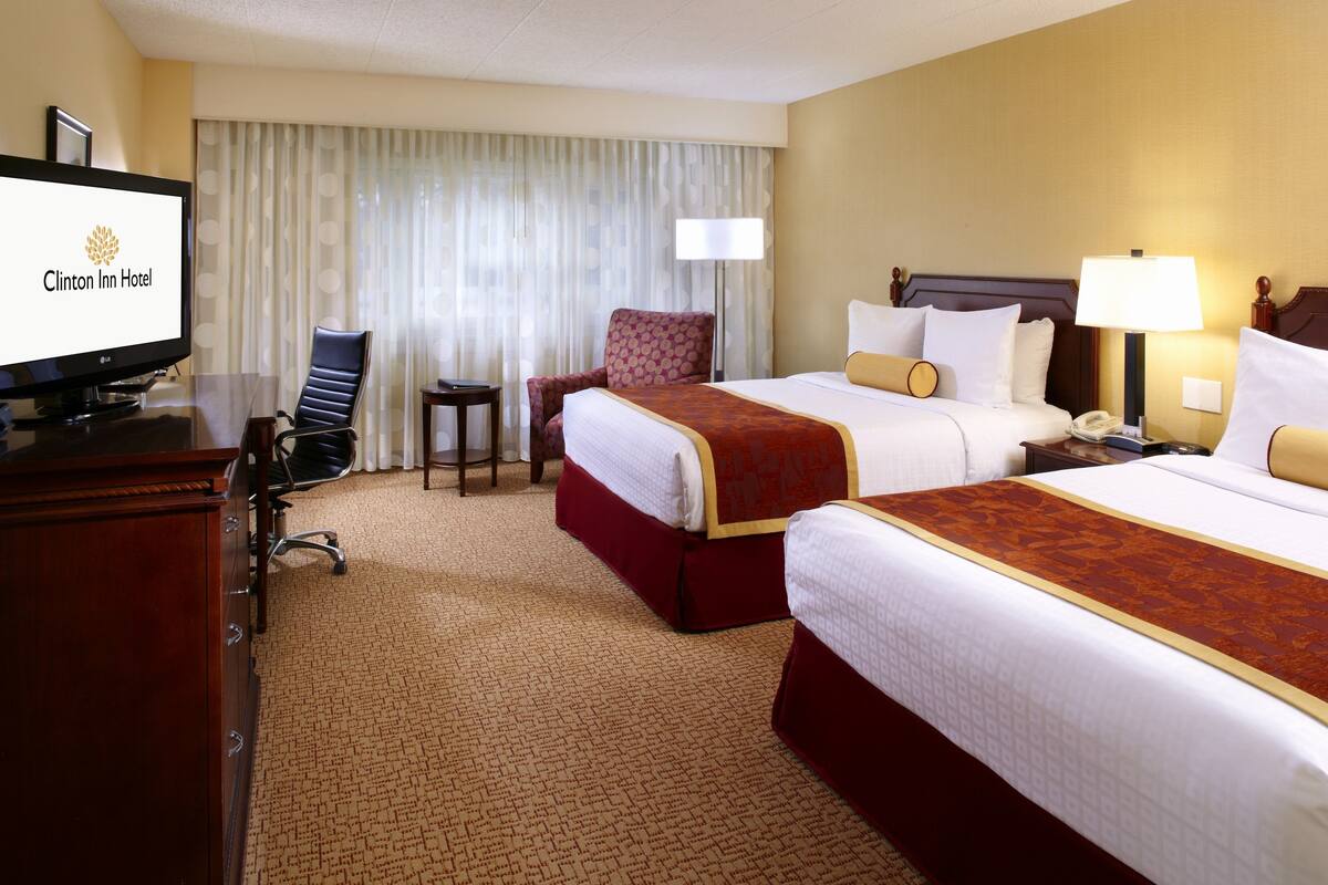A hotel room with two beds, a desk with a television, a chair, and a lamp.