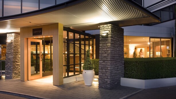 A modern hotel entrance with a stone facade, large glass doors, and a well-lit interior visible through the windows.