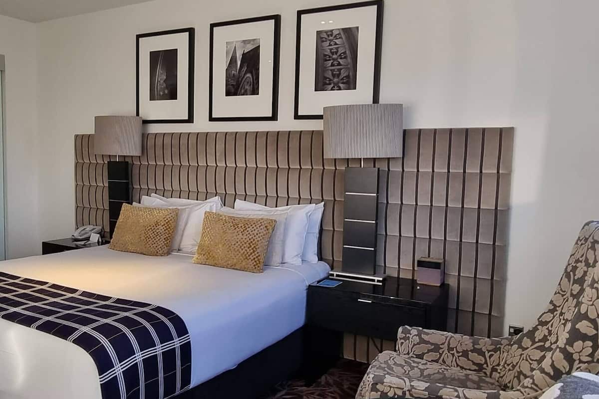 A hotel room with a large bed, a nightstand, a lamp, a chair, and framed pictures on the wall.