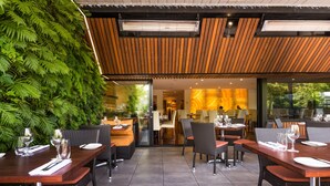 An outdoor dining area with wooden tables and chairs, a green wall, and a view of the restaurant interior.