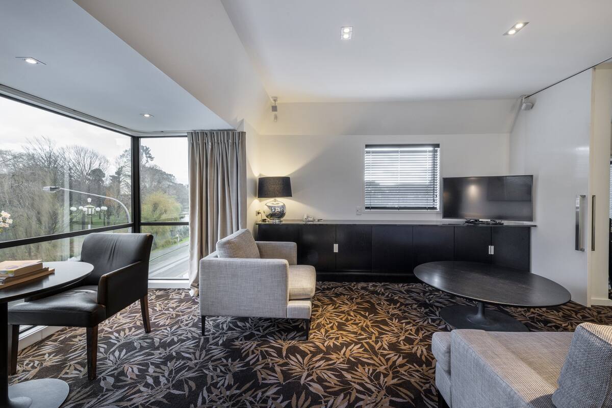 A modern hotel room with a large window, a sofa, a dining table, a TV, and a patterned carpet.