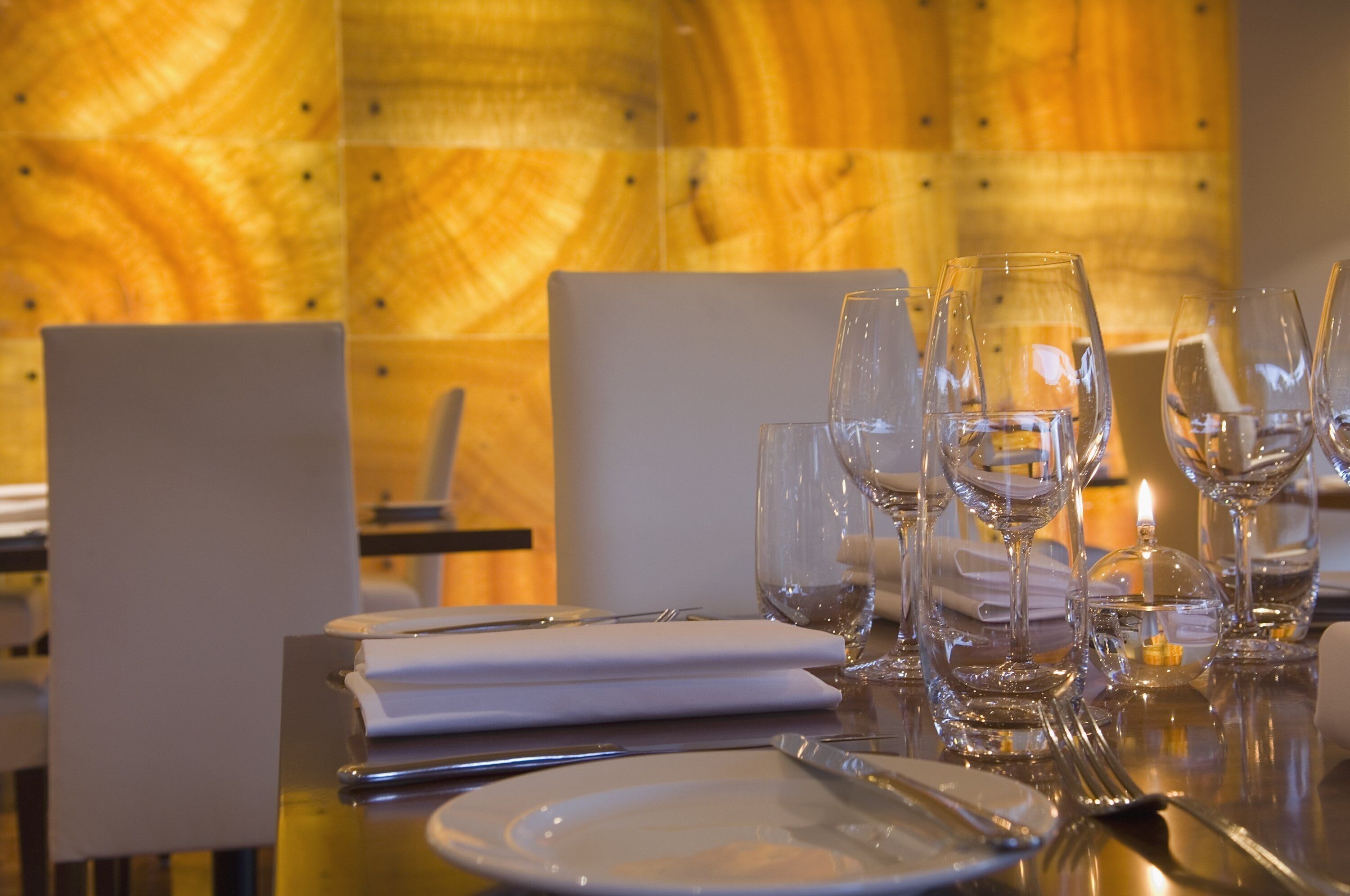 A dining table set with white plates, cutlery, and multiple wine glasses, with a candle and a menu on the table.