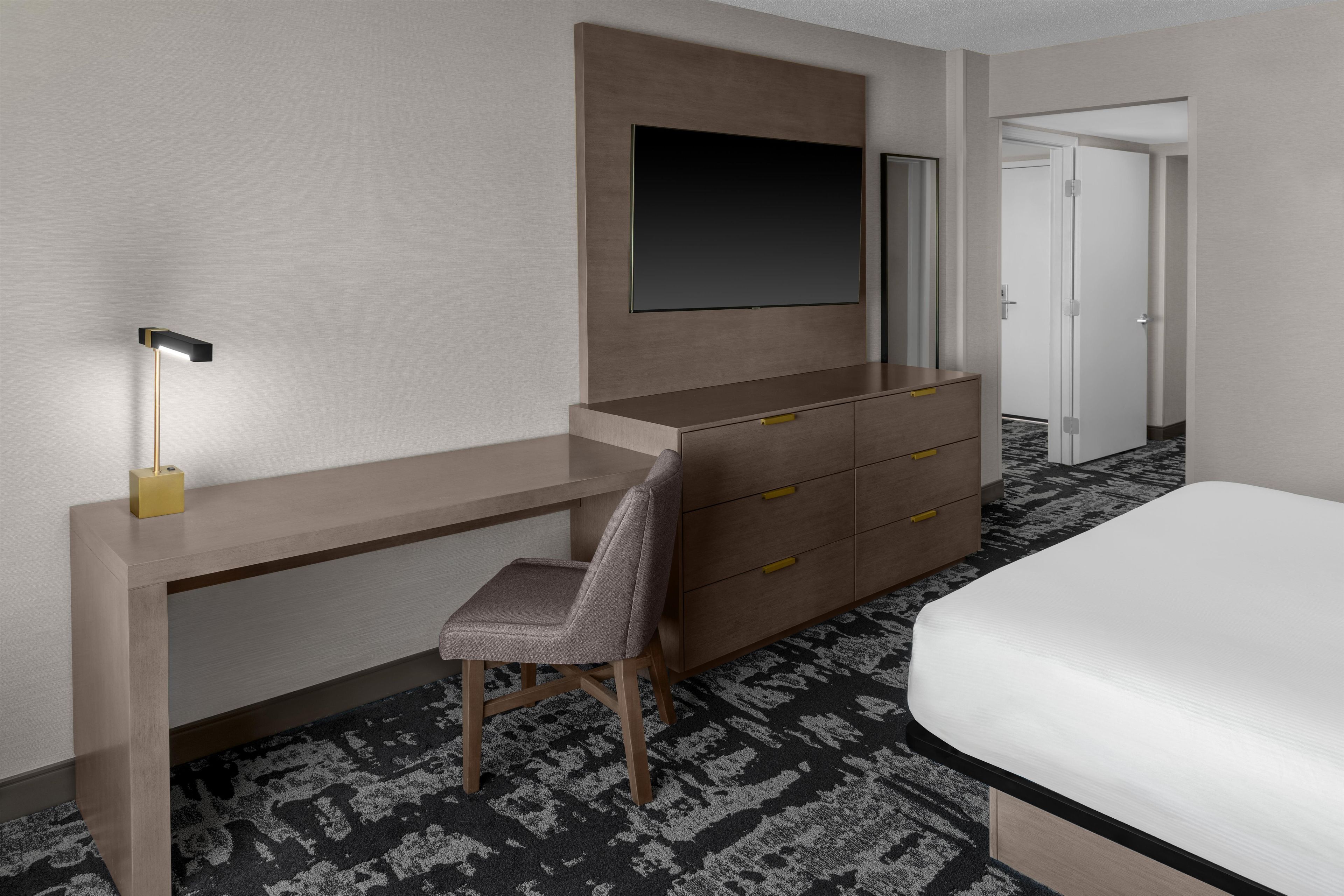 A hotel room with a bed, a desk with a chair, a dresser, and a flat-screen TV mounted on the wall.