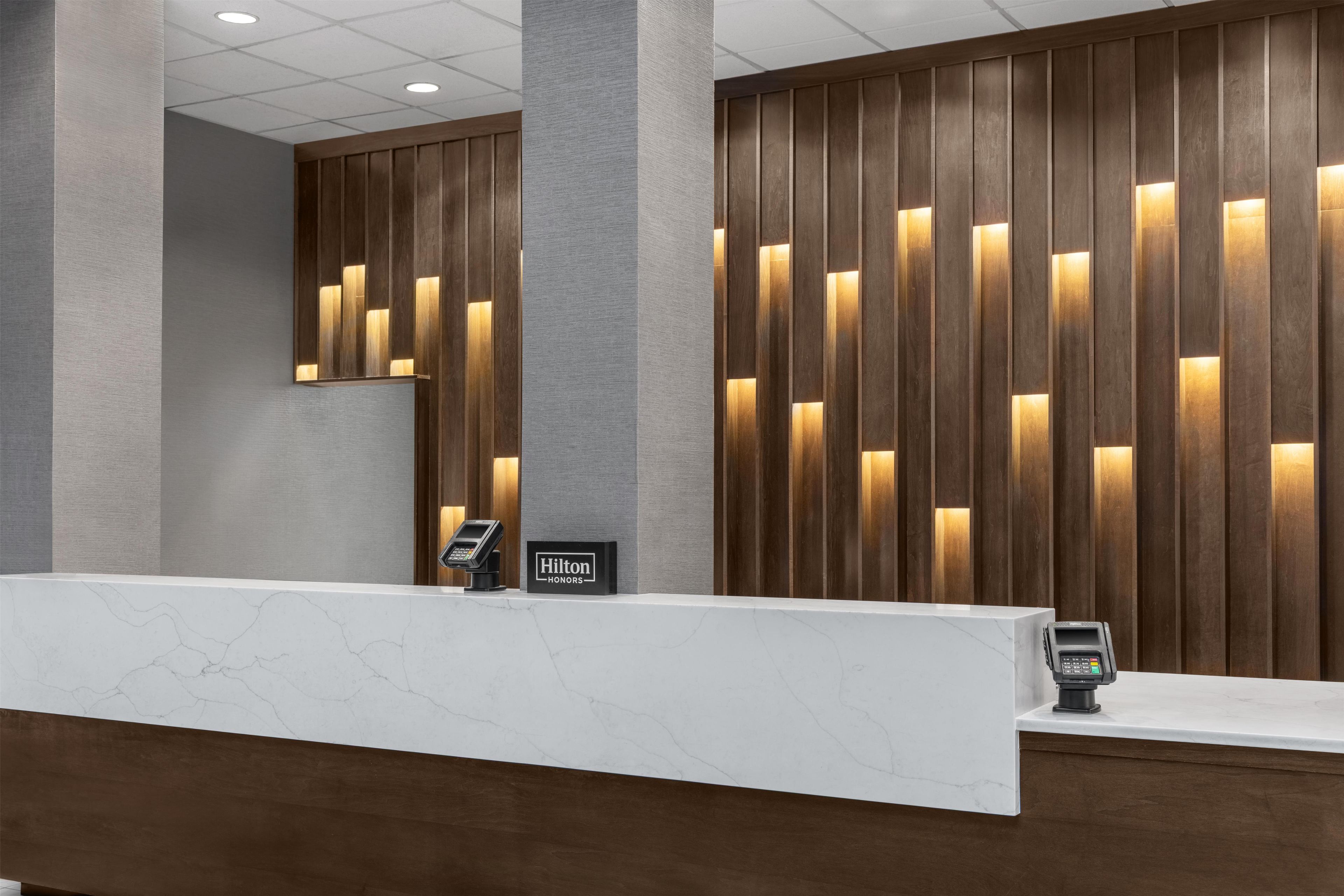 Ein Hotelrezeptionstresen mit Marmorplatte, einem digitalen Bezahlsystem und einem Schild mit der Aufschrift 'Hilton Honors'.