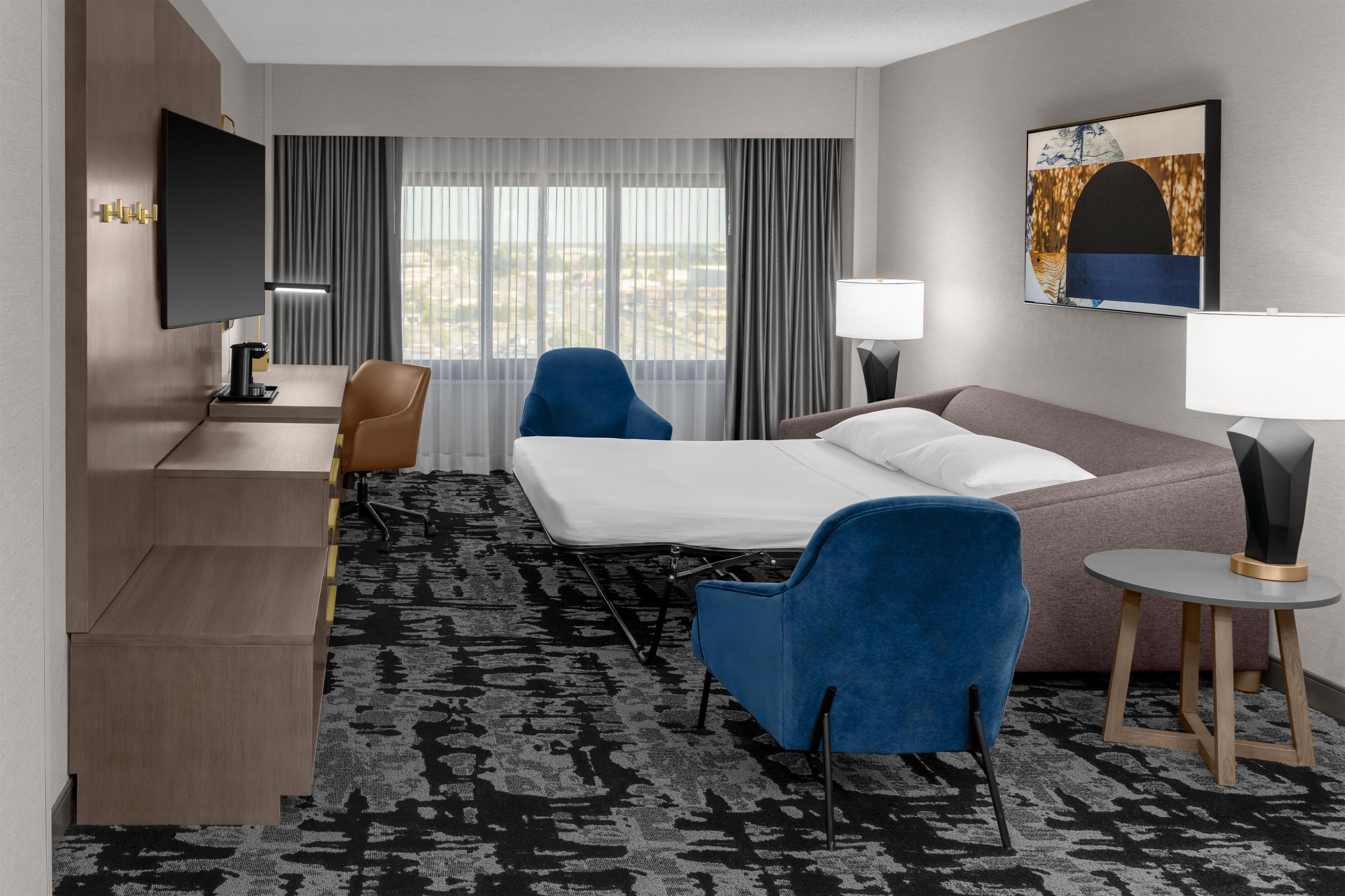 A hotel room with a bed, two chairs, a desk with a TV, a lamp, and a painting on the wall.