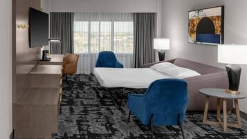 A hotel room with a bed, two chairs, a desk with a TV, a lamp, and a painting on the wall.