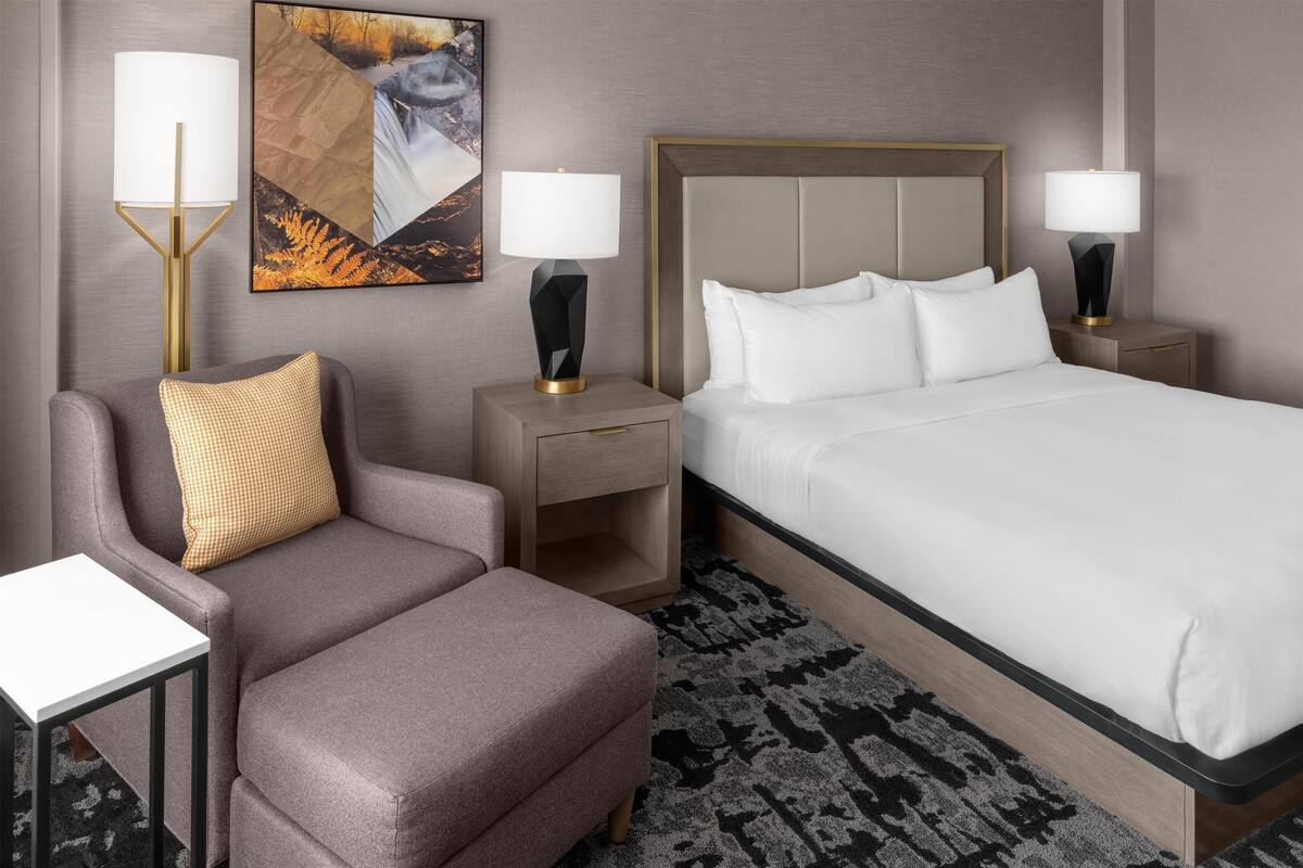 Habitación de hotel con una cama, un sofá, dos mesitas de noche con lámparas, una pequeña mesa y un cuadro colgado en la pared.
