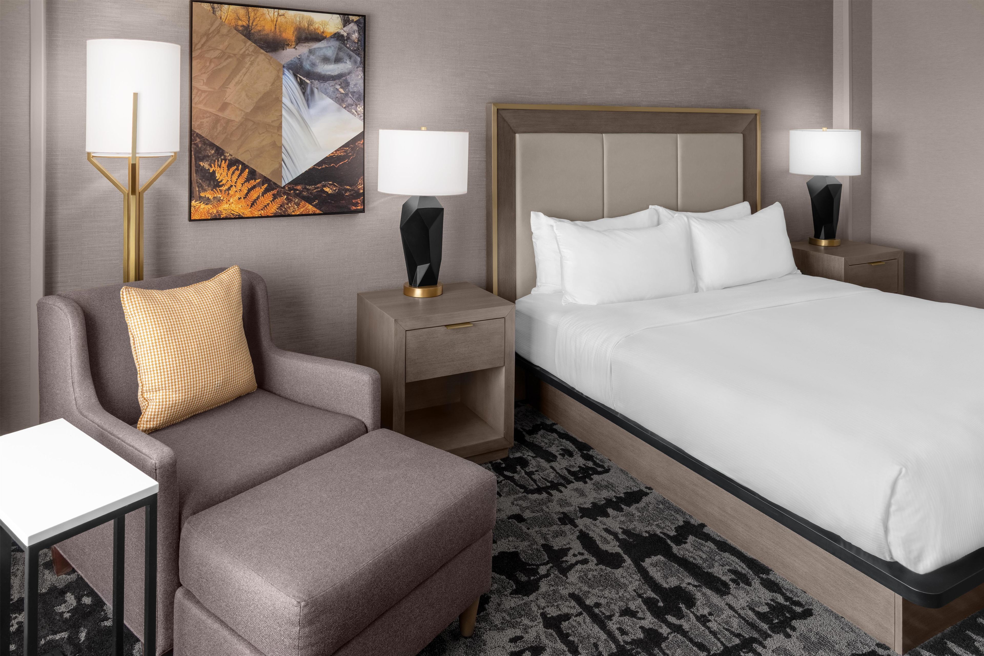 A hotel room with a bed, two nightstands, a chair, a desk, and a wall-mounted lamp.