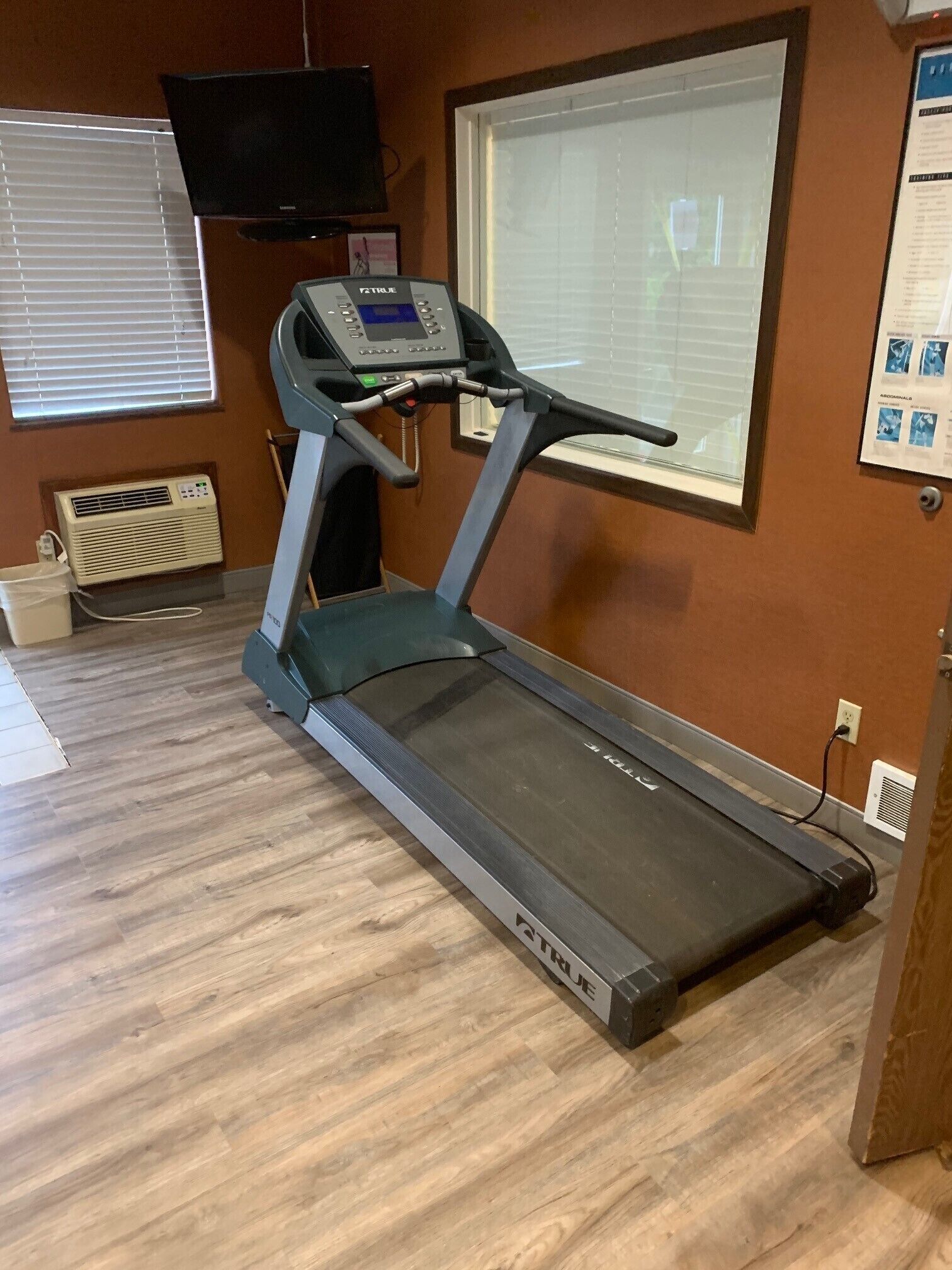 A treadmill in a room with wooden flooring, a wall-mounted TV, and a window with blinds.
