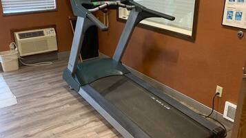 A treadmill in a room with wooden flooring, a wall-mounted TV, and a window with blinds.