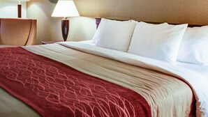 A hotel room with a neatly made bed, two bedside lamps, a round mirror on the wall, and a red patterned throw blanket.