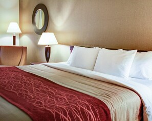 A hotel room with a neatly made bed, two bedside lamps, a round mirror on the wall, and a red patterned throw blanket.