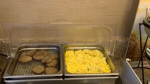 A buffet setup with two trays of food, one containing cookies and the other scrambled eggs, and two metal serving spoons on a granite countertop.