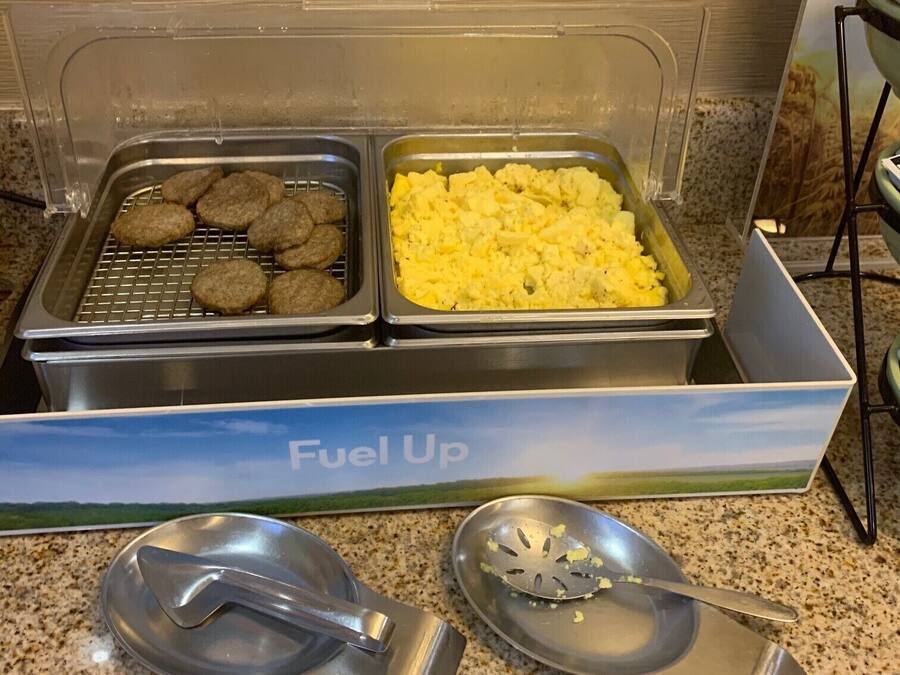 A buffet setup with two trays of food, one containing cookies and the other scrambled eggs, and two metal serving spoons on a granite countertop.