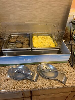 A buffet setup with two trays of food, one containing cookies and the other scrambled eggs, and two metal serving spoons on a granite countertop.