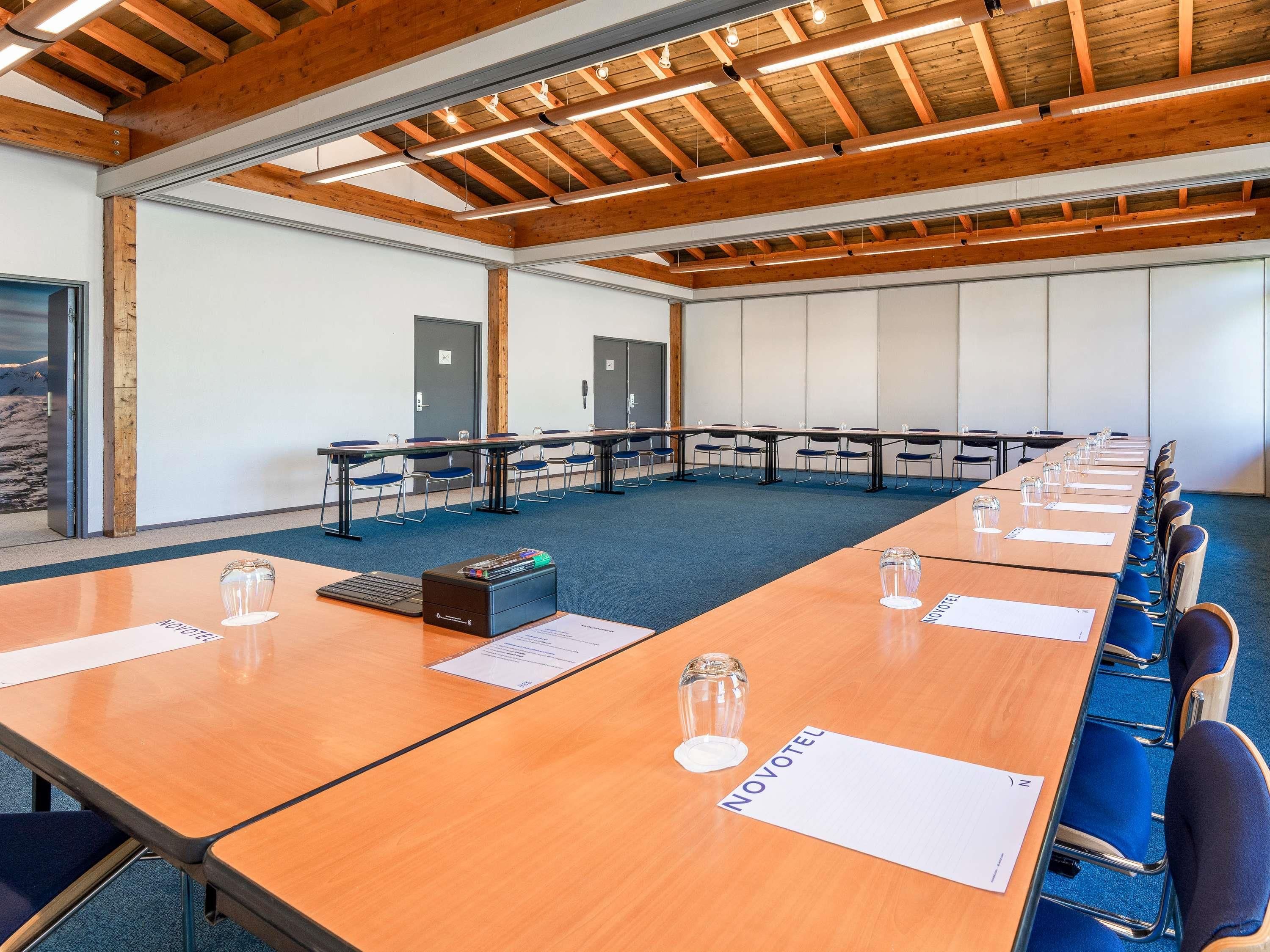 Un salón de conferencias con vigas de madera, mesas con sillas y vistas a montañas a través de una puerta.