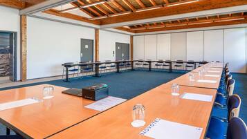 Un salón de conferencias con vigas de madera, mesas con sillas y vistas a montañas a través de una puerta.