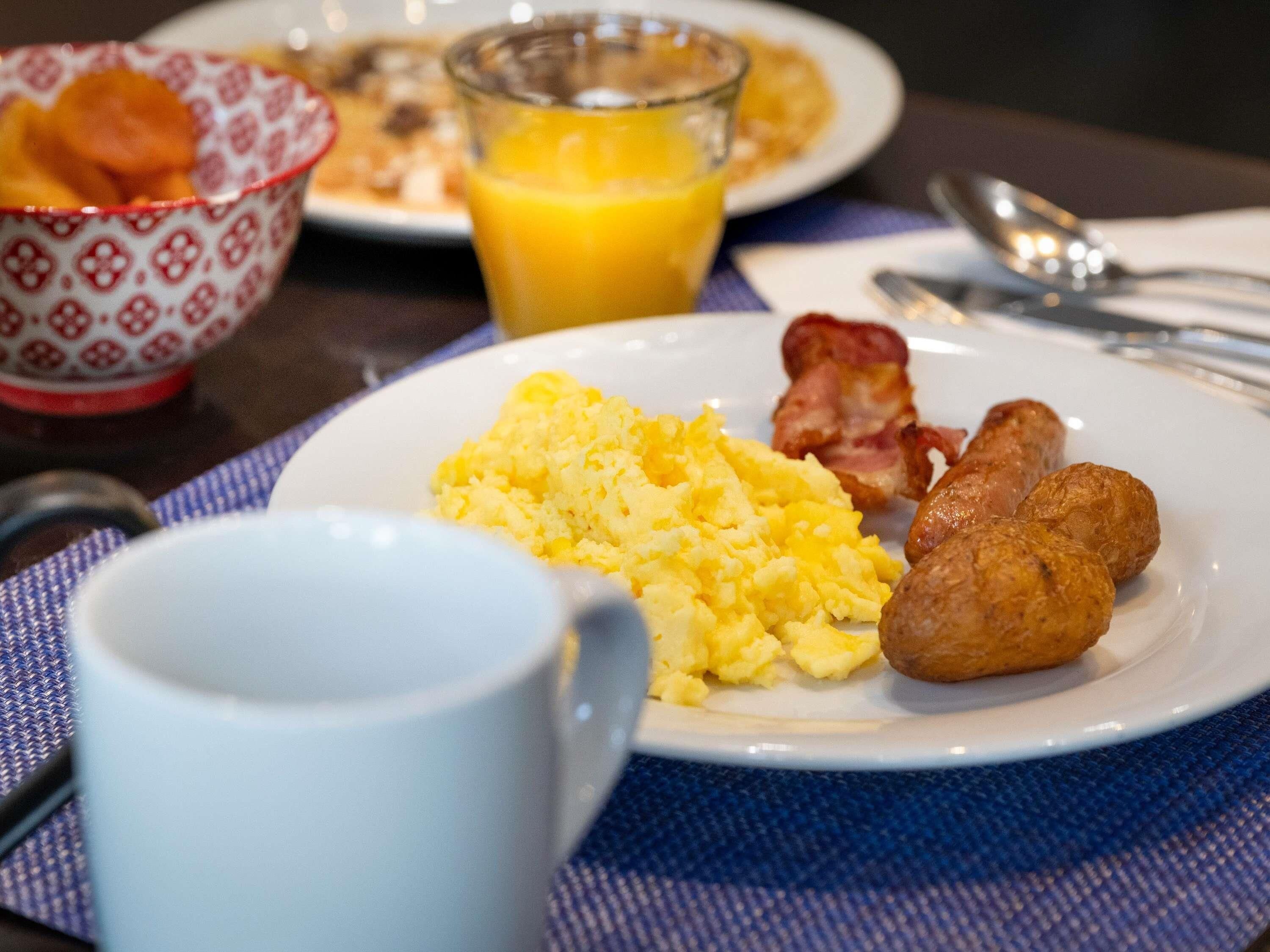 Ein Frühstücksteller mit Rührei, Speck und Würstchen, ein Glas Orangensaft und eine Kaffeetasse auf einem Tisch mit einer blauen Tischmatte.