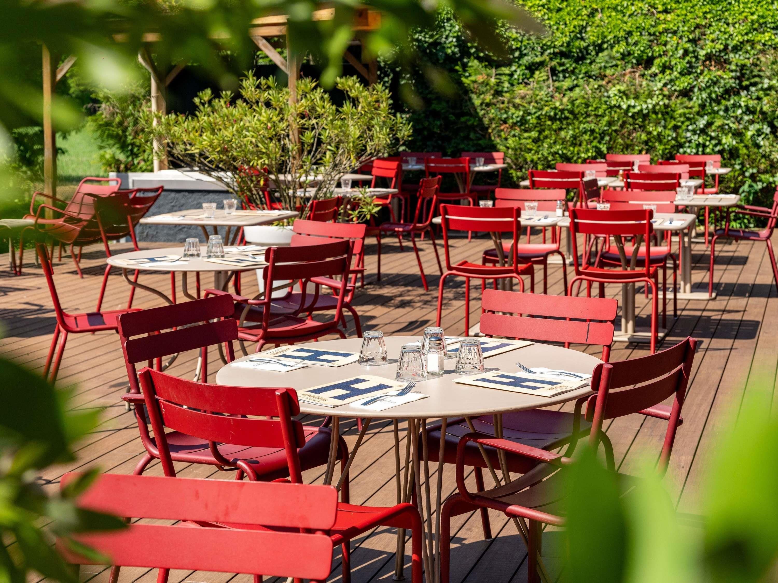 An outdoor dining area with red chairs and tables, surrounded by greenery.