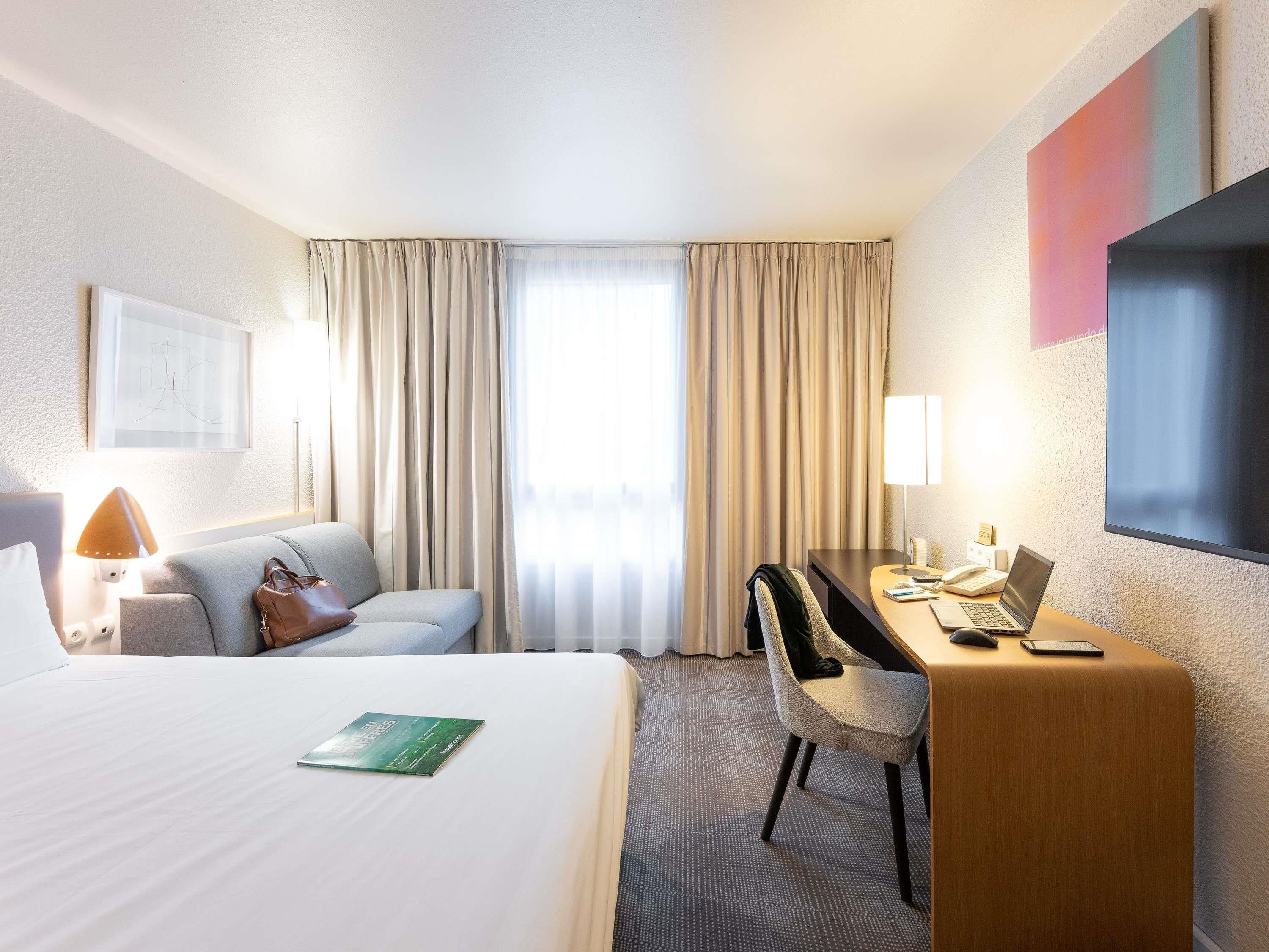 A hotel room with a bed, a desk with a laptop, a chair, a lamp, a handbag, and a window with curtains.