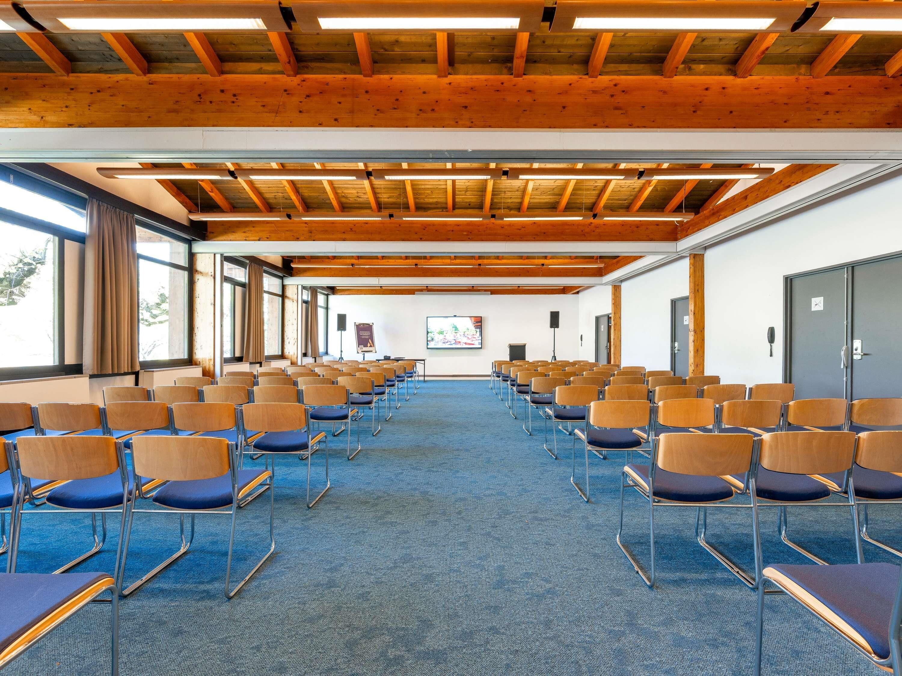 Een vergaderzaal met rijen stoelen, een projectiescherm en een houten plafond.