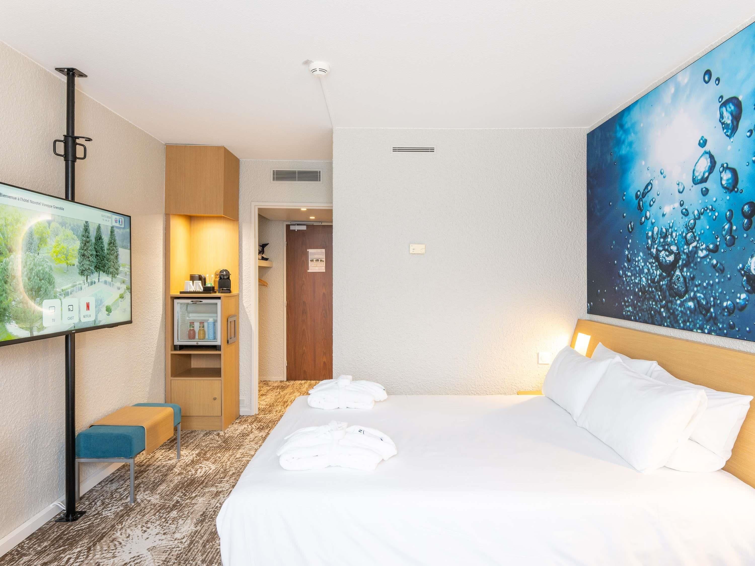 A hotel room with a bed, a TV, a desk with a computer, and a painting on the wall.