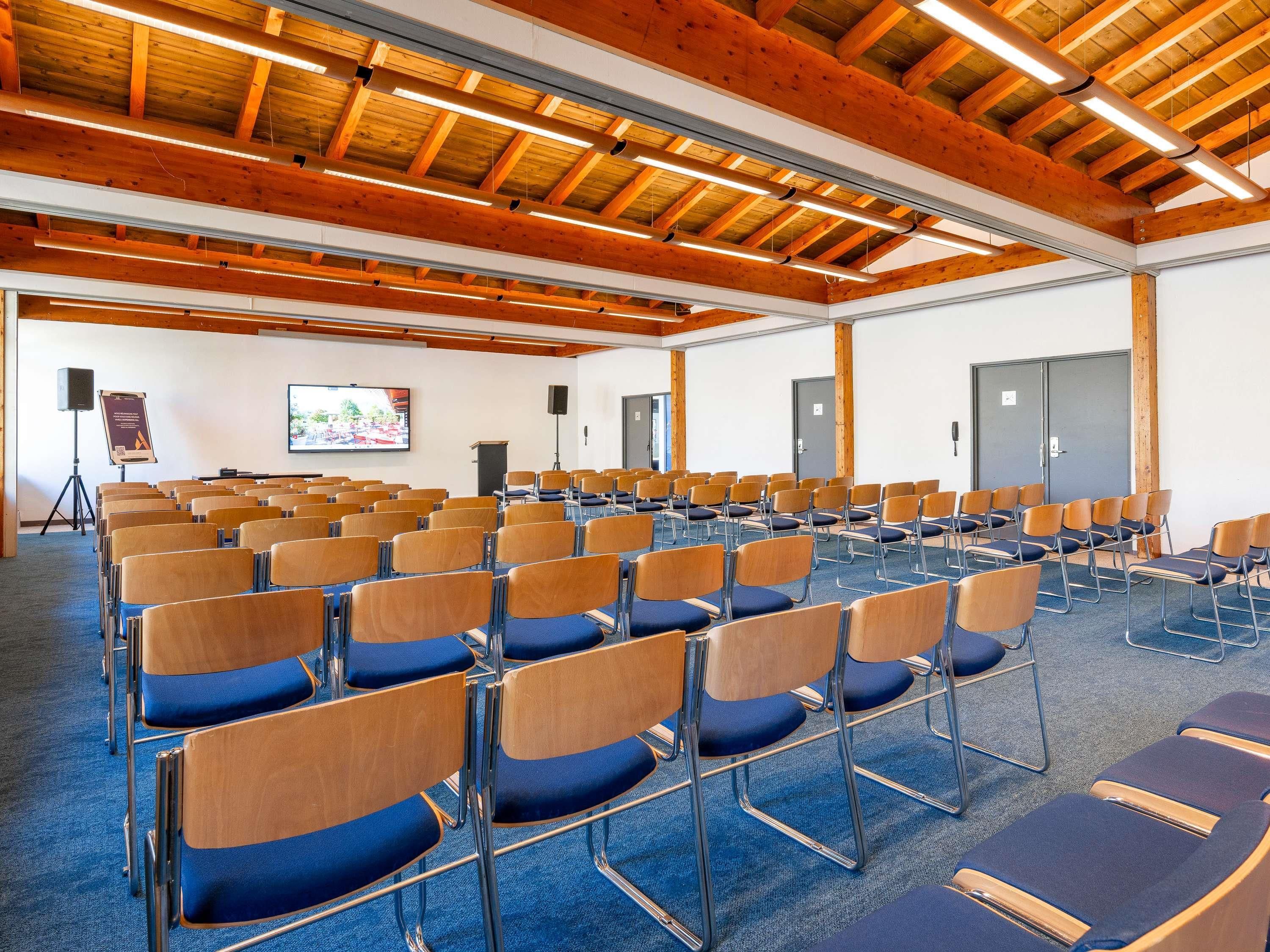 Un salón de conferencias con filas de sillas, una pantalla de proyector y un orador.
