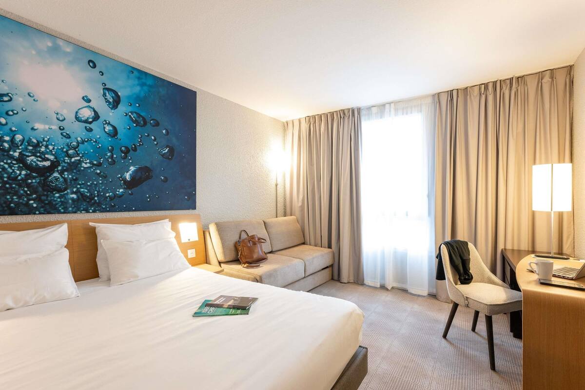 Une chambre d’hôtel avec un grand lit, un bureau avec une chaise, un canapé et une grande fenêtre avec des rideaux.