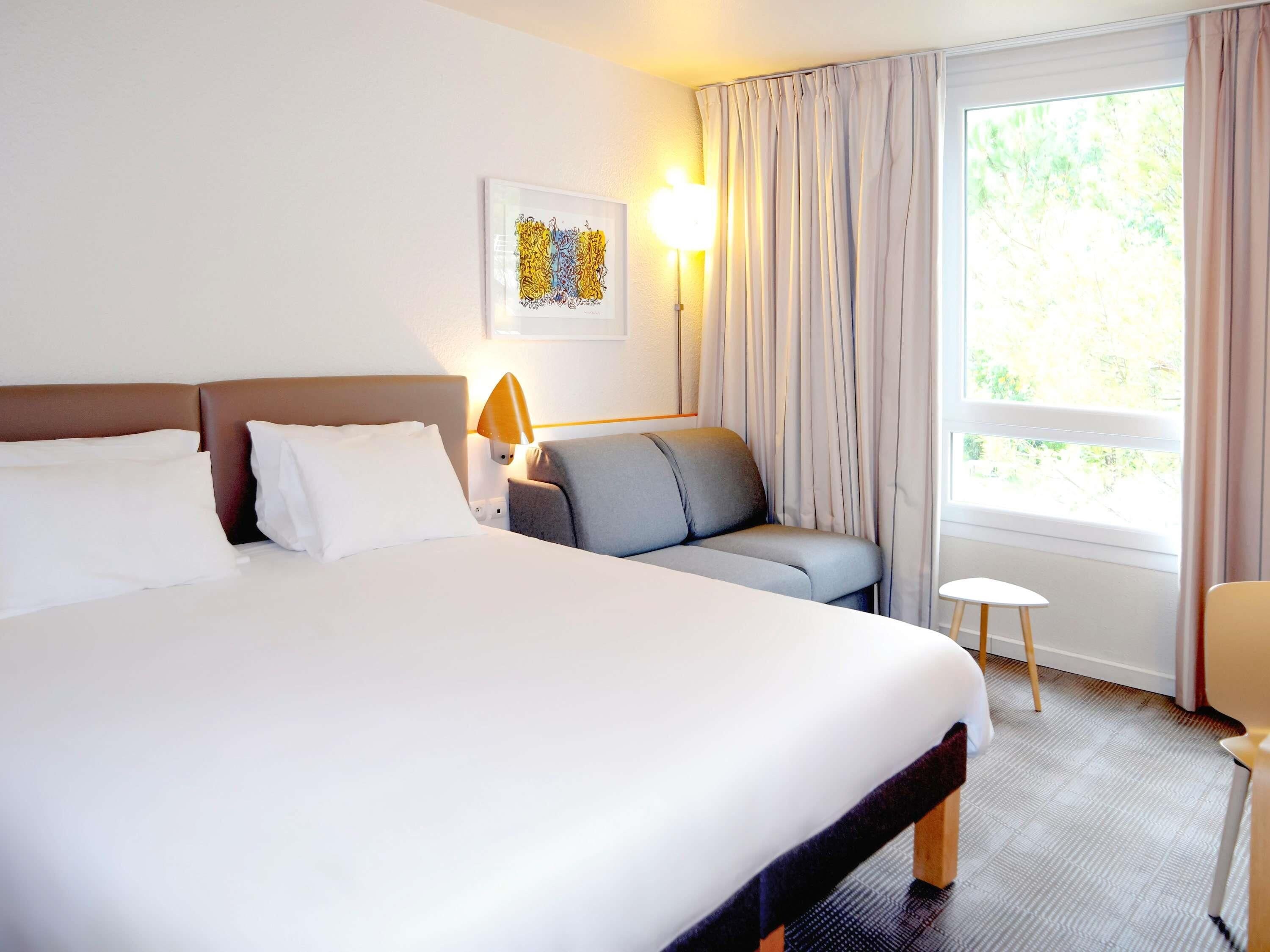 Ein Hotelzimmer mit einem Bett, einem grauen Sofa, einem kleinen Tisch, einer Lampe und einem Fenster mit Vorhängen.