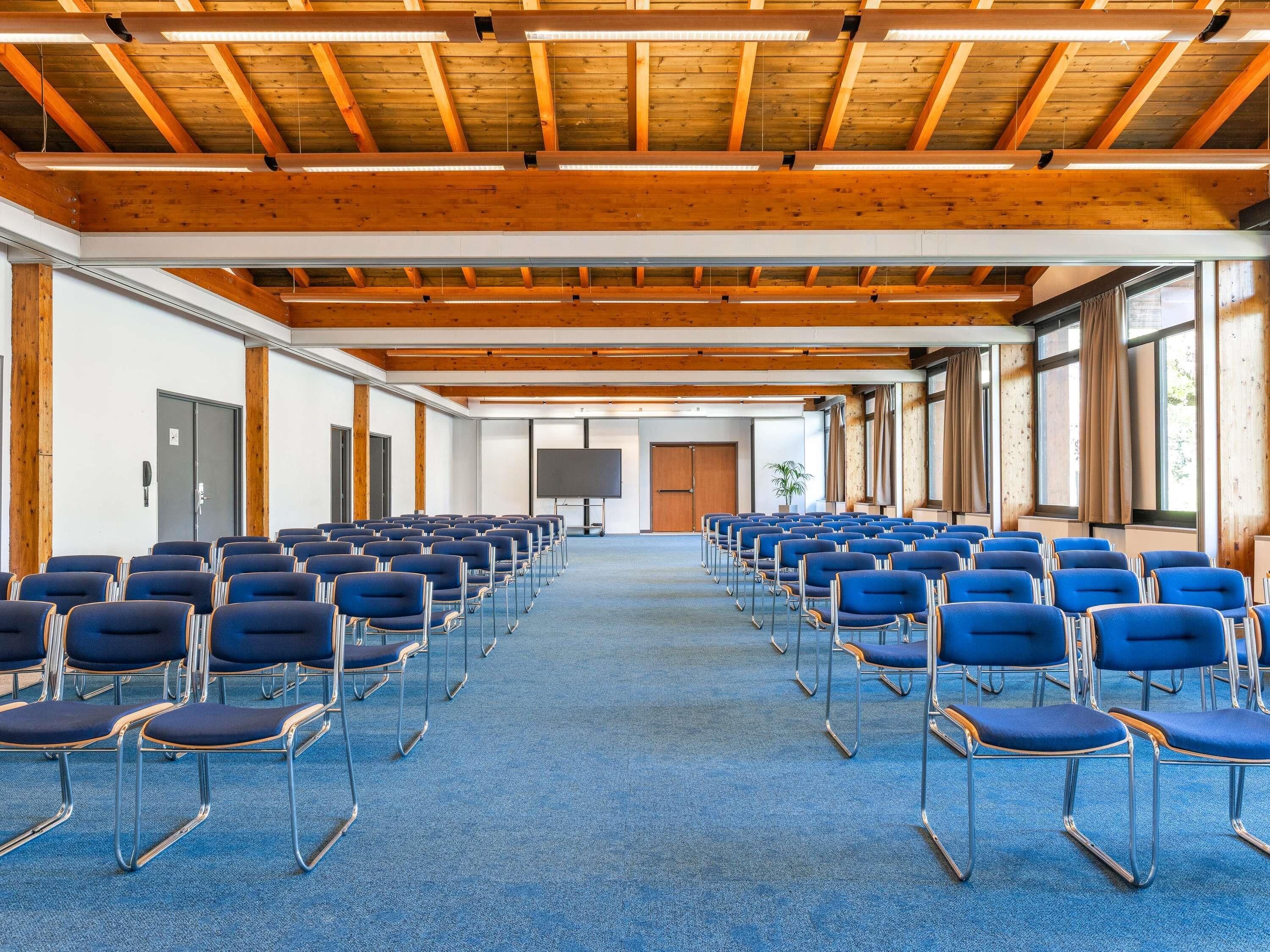 Een congreszaal met rijen stoelen, houten balken in het plafond en grote ramen.