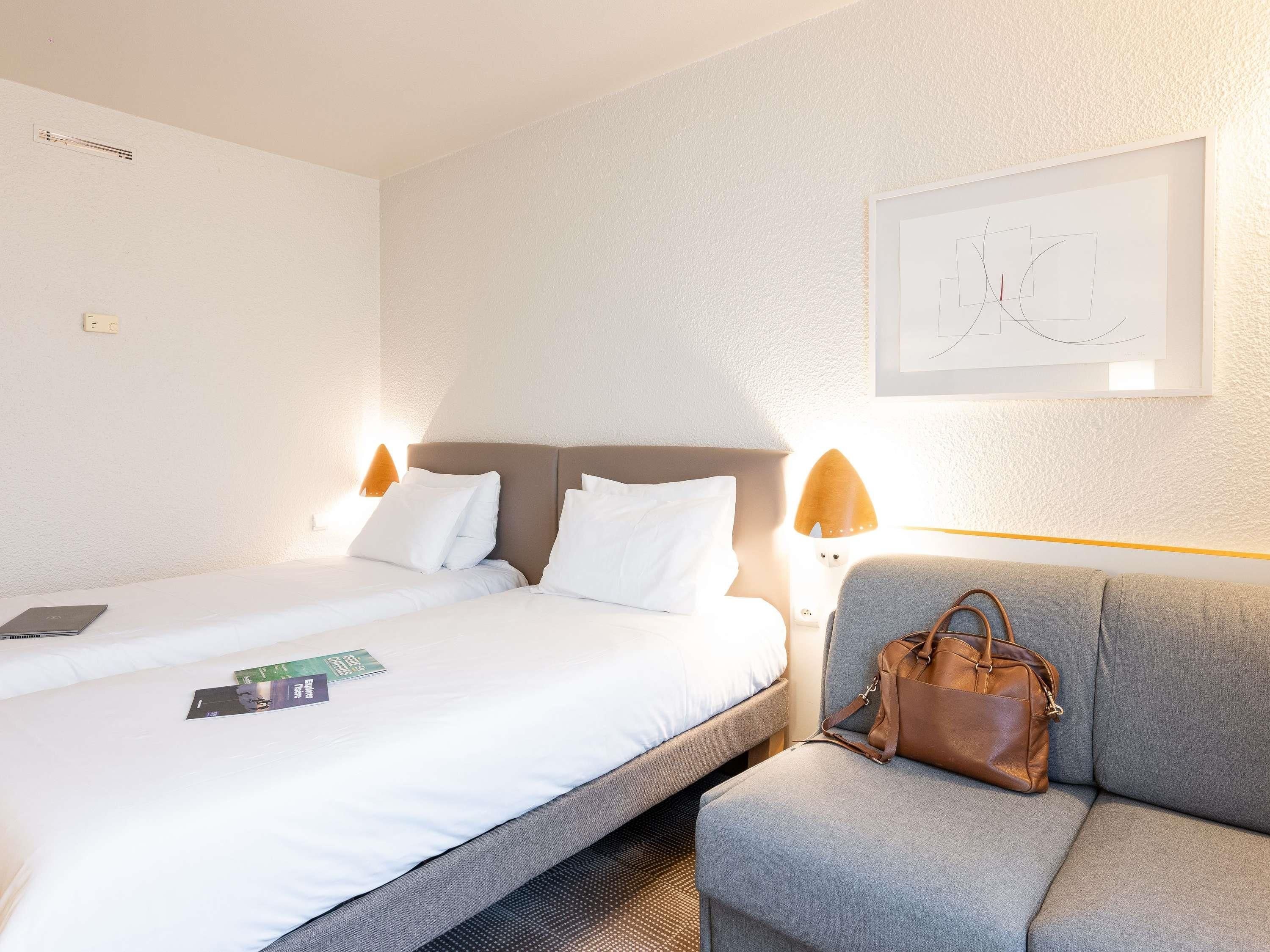 Een hotelkamer met een bed, een grijze fauteuil, een bruine handtas en een lijst met abstracte kunst aan de muur.
