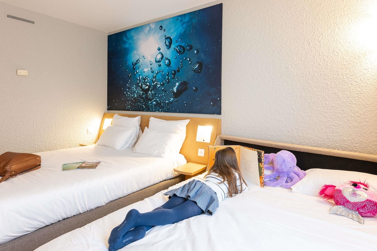 Une chambre d’hôtel avec deux lits, une grande peinture représentant des gouttes d’eau sur le mur, et une personne allongée sur l’un des lits, en train de lire un livre.