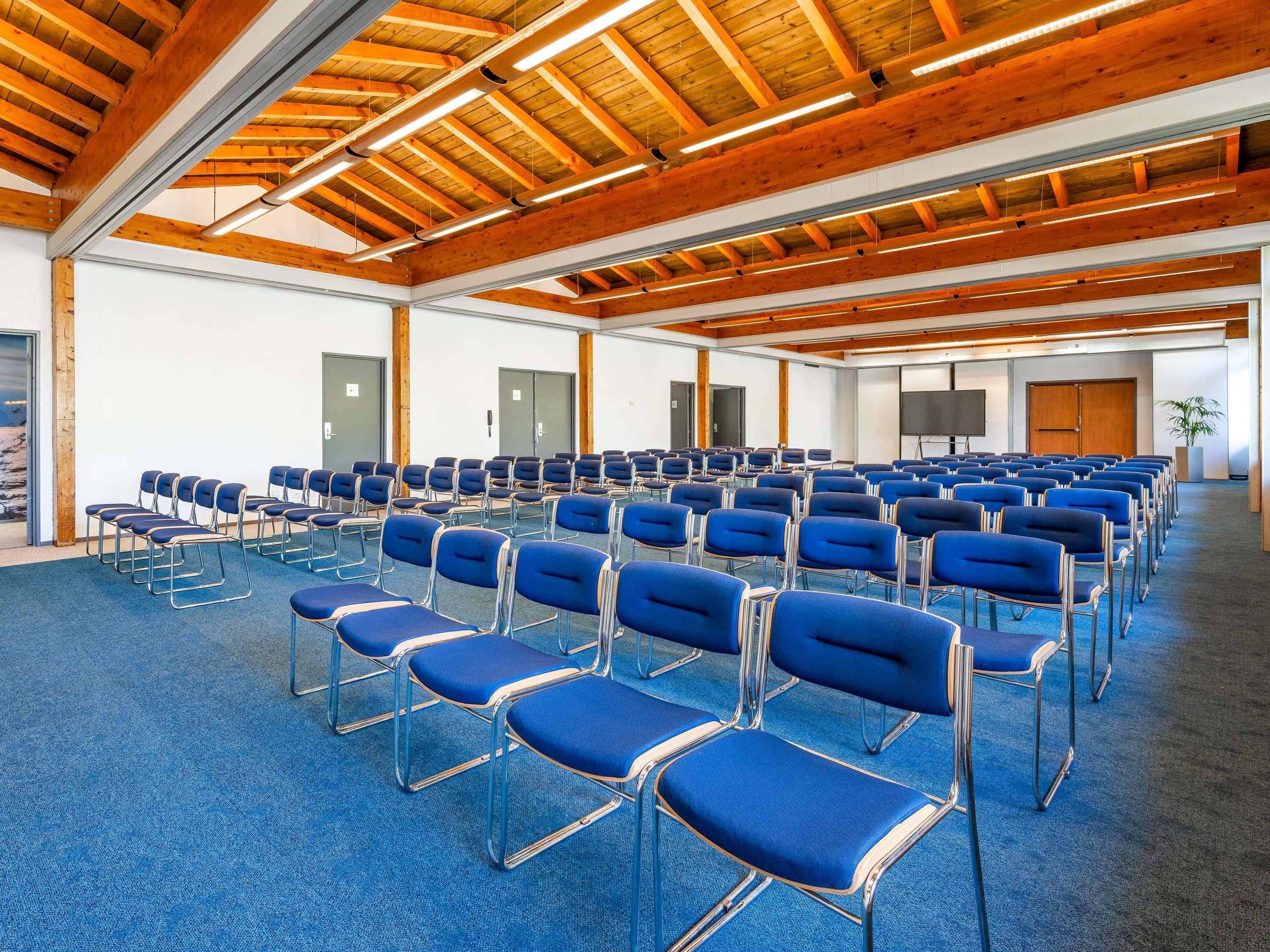 Una sala de conferències amb files de cadires blaves, bigues de sostre de fusta i una pantalla gran.