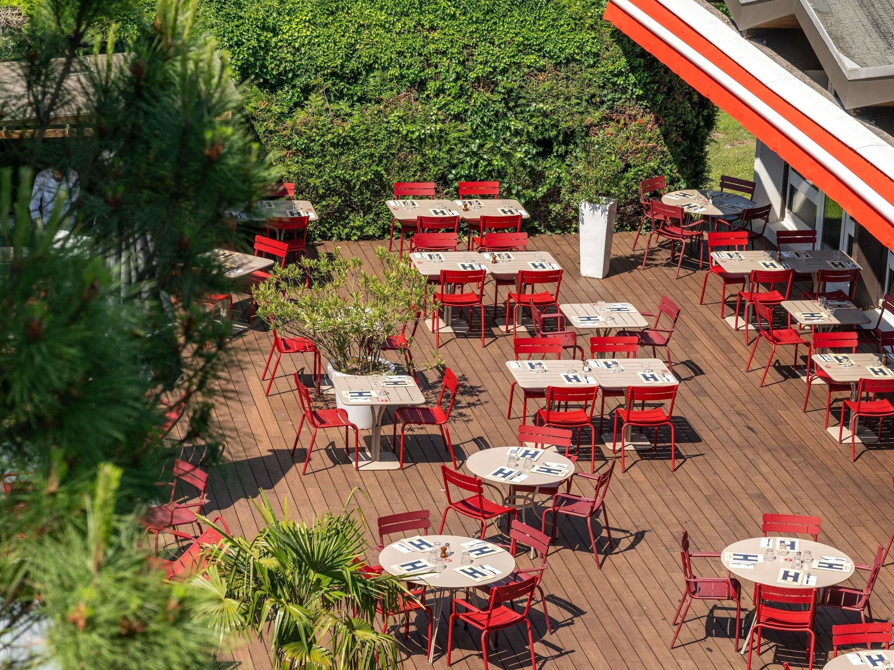 An outdoor patio with multiple tables and chairs, surrounded by greenery and a building with an orange and white facade.
