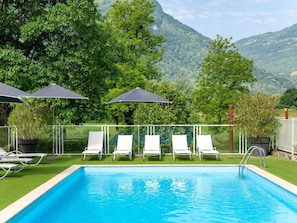 Piscina con sillas de descanso y sombrillas, rodeada de vegetación y montañas.