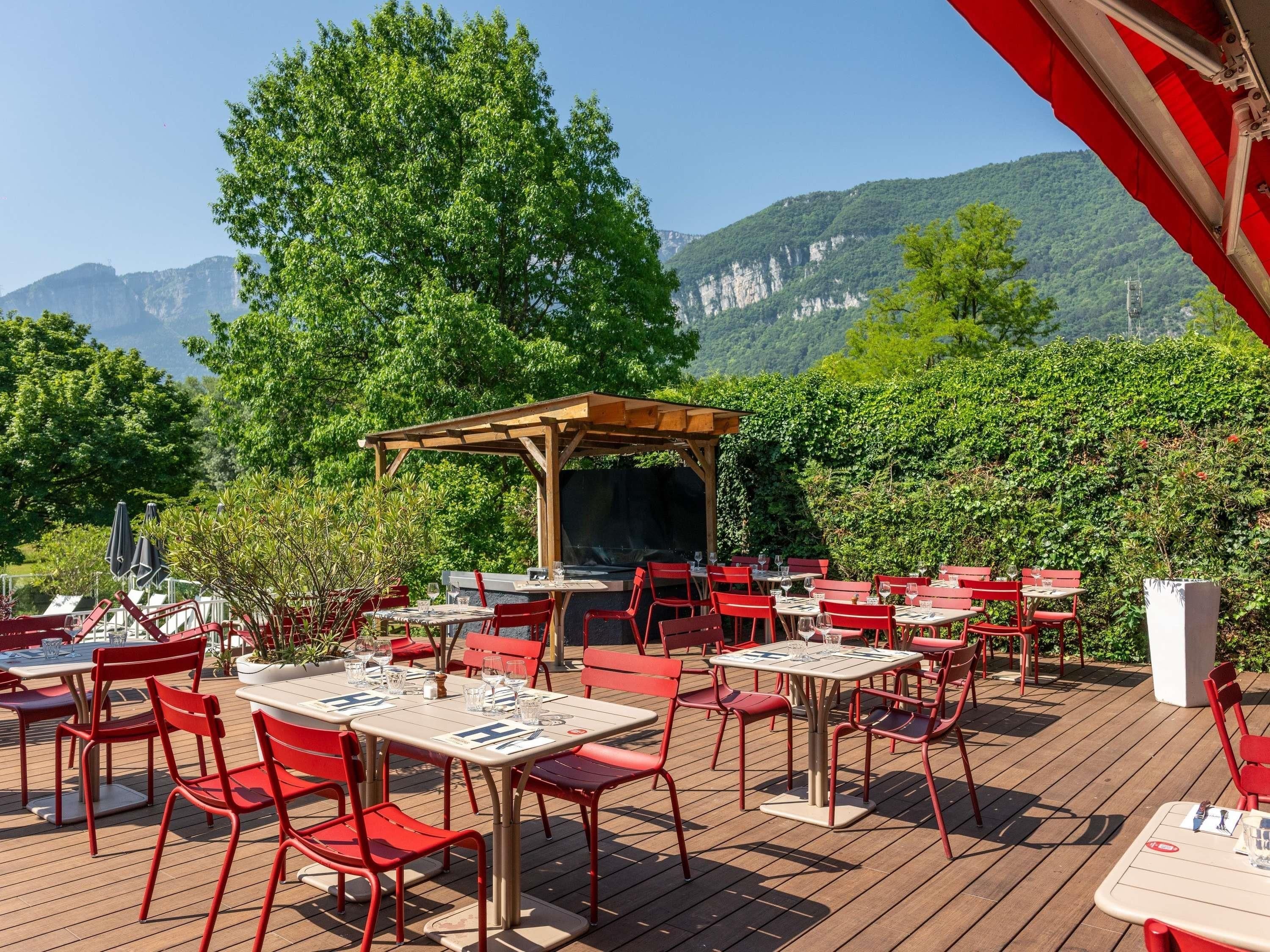 Un espace extérieur aménagé pour les repas, avec des chaises et des tables rouges, une pergola en bois et une vue sur les montagnes.