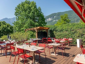 An outdoor dining area with red chairs and tables, a wooden pergola, and a view of mountains. - Novotel Grenoble Nord Voreppe (Voreppe)