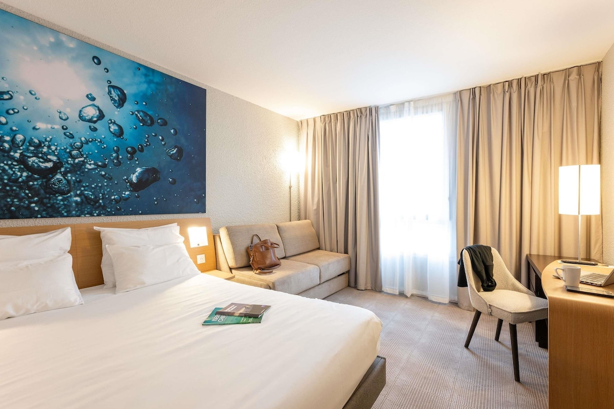Een hotelkamer met een groot bed, een bureau met stoel, een bank en een groot raam met gordijnen.