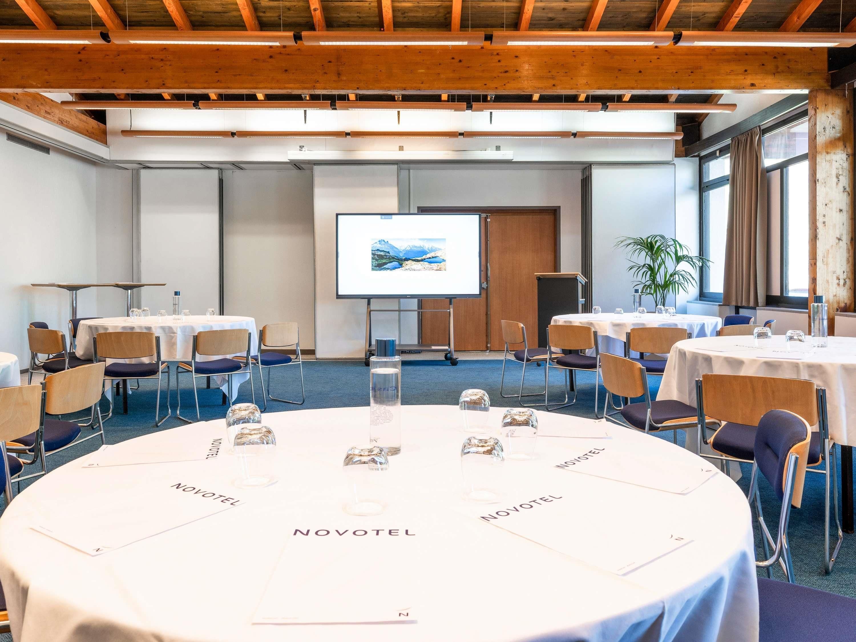 Un salón de conferencias con mesas redondas, sillas, una pantalla de proyector y techo de madera.