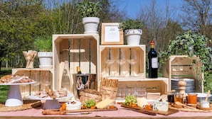 Un coin pique-nique aménagé en plein air avec des caisses en bois, une bouteille de vin, ainsi que divers fromages et fruits.