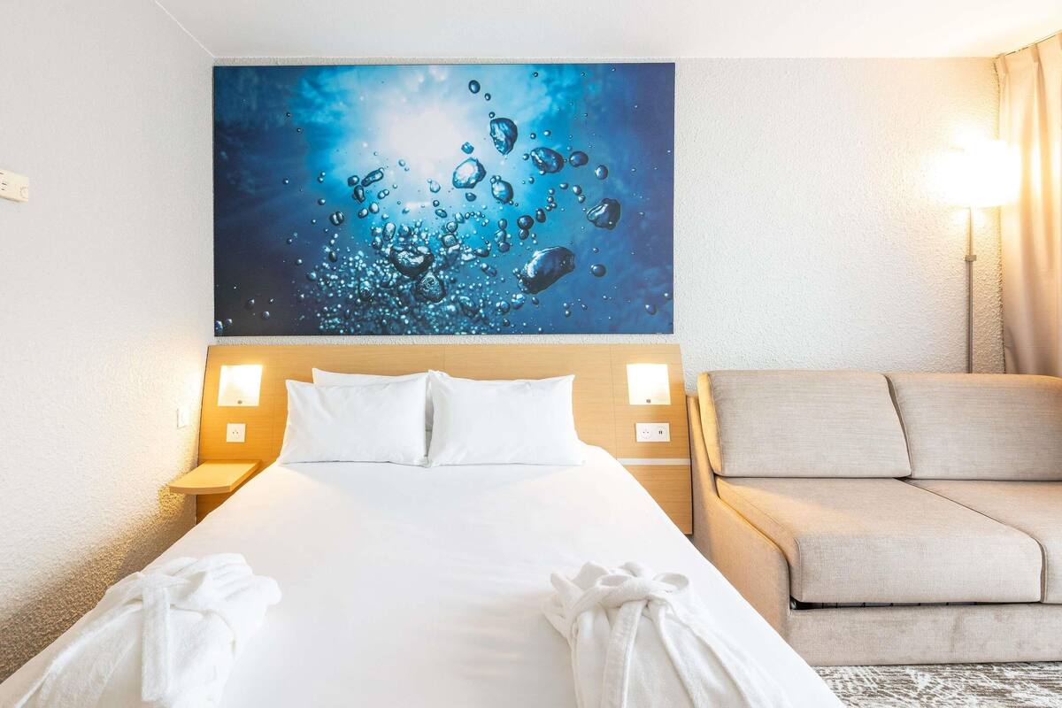 Une chambre d’hôtel avec un grand lit, deux lampes de chevet, un canapé beige et un tableau représentant des bulles d’eau sur le mur.