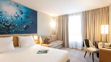 A hotel room with a large bed, a desk with a chair, a sofa, and a large window with curtains.